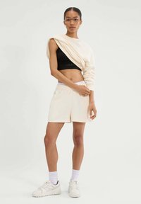 Kremowy sweatshirt nałożony na czarny top, w połączeniu z jasnymi szortami. Model nosi białe sneakersy i skarpetki sięgające kostek, oraz przezroczyste okulary.