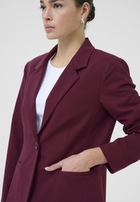 Burgundský blejzr s přiléhavým střihem, klopami, jednobuttonovým zapínáním a bočními kapsami. Hladká, strukturovaná tkanina a dlouhé rukávy.