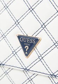 Borsa bianca con un motivo a rombi cucito con filo blu. Una placca triangolare in blu navy con dettagli dorati è posizionata al centro.