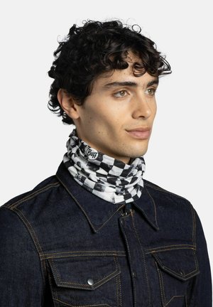 Giovane uomo con capelli ricci che indossa un passamontagna a scacchi bianco e nero e una giacca di jeans scura con cuciture gialle.