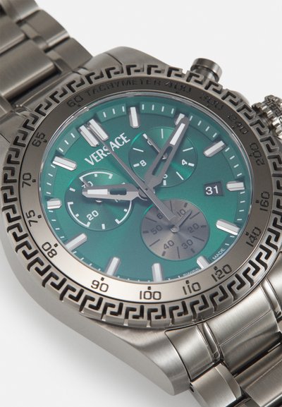 Silver chronograph watch with a green dial, stainless steel case, intricate patterned bezel, and three sub-dials. Date display at 4 o'clock.
