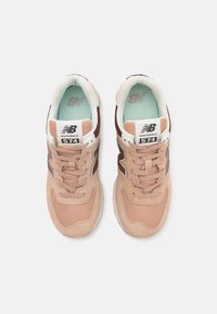 Sneakers New Balance 574 w kolorze brzoskwiniowym i brązowym, wykonane z zamszu i siateczki, z wyściełanym kołnierzem, sznurowadłami oraz akcentami z logo.