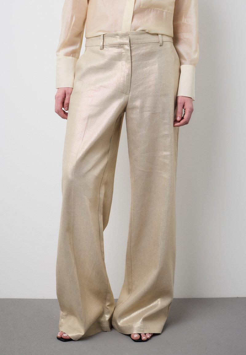 Beige linnen broek met brede pijpen en glans, gecombineerd met een crèmekleurig transparant blouse en zwarte sandalen met open tenen, op een grijze vloer.