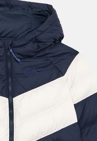 Navy and cream puffer jacket with a hood, featuring diagonal color-blocking and an embroidered logo on the chest, textured fabric.