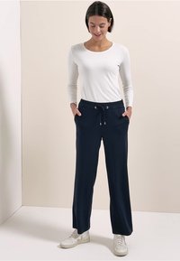 Nainen seisoo käsineet taskuissa, pukeutuneena valkoiseen pitkähihainen puseroon, tummansinisiin naruvetoketjuhousuihin ja valkoisiin tennareihin yksiväristä taustaa vasten.