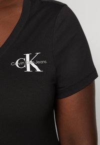 Čierne bavlnené tričko s V-výstrihom, ktoré má na hrudi biele logo "CK" a text "Calvin Klein Jeans". Krátke rukávy a hladká textúra.