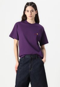Purple cotton t-shirt with a round neckline and short sleeves, featuring a small embroidered logo in red. Worn with dark denim trousers.