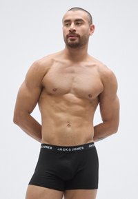 Svarta boxerbriefs med ett elastiskt midjeband med varumärkeslogga. Modellen har en muskulös kroppsbyggnad, kort hår och skägg, och står mot en ljus bakgrund.