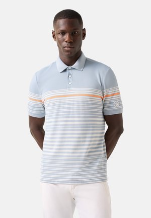 Jonge man staat voorover gericht, draagt een lichtblauw poloshirt met oranje en witte horizontale strepen en een witte broek, handen achter de rug.