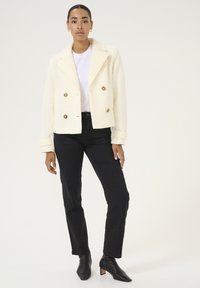 Veste double boutonnage crème avec des boutons dorés, tissu texturé, large col et manches retroussées, associée à un jean skinny noir et des bottines.