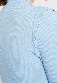 Hellblauer gerippter Pullover mit hohem Kragen und einzigartigem weißen Schlaufenakzent entlang der Schulternaht. Weicher Stoff und taillierte Passform.