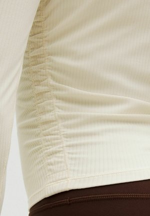 Primo piano di un top beige a maniche lunghe a coste con cucitura laterale arricciata indossato sopra pantaloni marrone scuro.