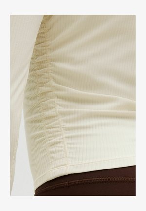 Primo piano di un top beige a maniche lunghe a coste con cucitura laterale arricciata indossato sopra pantaloni marrone scuro.