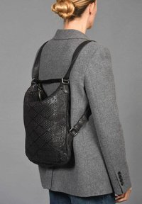 Zwarte geweven leren rugzak met verstelbare schouderbanden, met een gladde textuur en een ruitpatroon, gedragen over een grijze blazer.