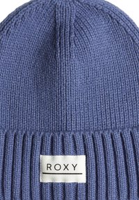 Blauwe gebreide beanie met geribbelde textuur, afgewerkt met een wit rechthoekig etiket met de merknaam "ROXY."