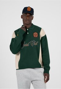 Polo verde e crema con logo ricamato sul lato sinistro del petto e scritta "Social Club". Indossato con pantaloni della tuta grigi e un cappellino abbinato.