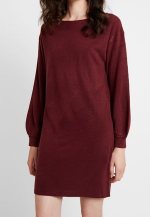 Femme portant une robe pull en maille bordeaux à manches longues avec poignets et ourlet côtelés sur un fond clair uni.
