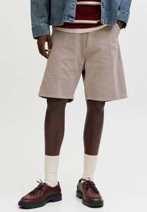 Oseba, oblečena v bež kratke hlače, krem nogavice, rjave usnjene čevlje, črtasto majico in jeans jakno, stoji ob enobarvnem ozadju.