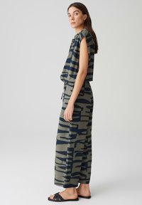 Abgeschnittenes, camoufliertes Oberteil in Navy und Grün, kombiniert mit weit geschnittenen Hosen mit einem ähnlichen Muster, ergänzt durch schwarze flache Sandalen.