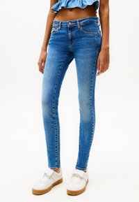 Kitsad sinised denim teksad keskmise kõrgusega vöökohaga, kerge kulumisega ja viie tasku disainiga, koos valgete tossudega, millel on kummi tald.