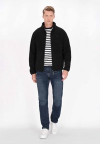 Giacca in pile nera con colletto alto, indossata sopra una maglietta a righe bianche e nere, abbinata a jeans blu e sneaker bianche.