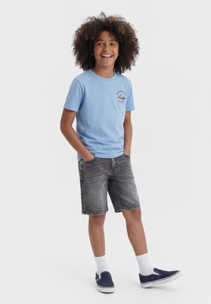 Niño sonriente con cabello rizado que lleva una camiseta azul claro con un pequeño gráfico, pantalones cortos de mezclilla grises, calcetines blancos y zapatos náuticos azul marino.
