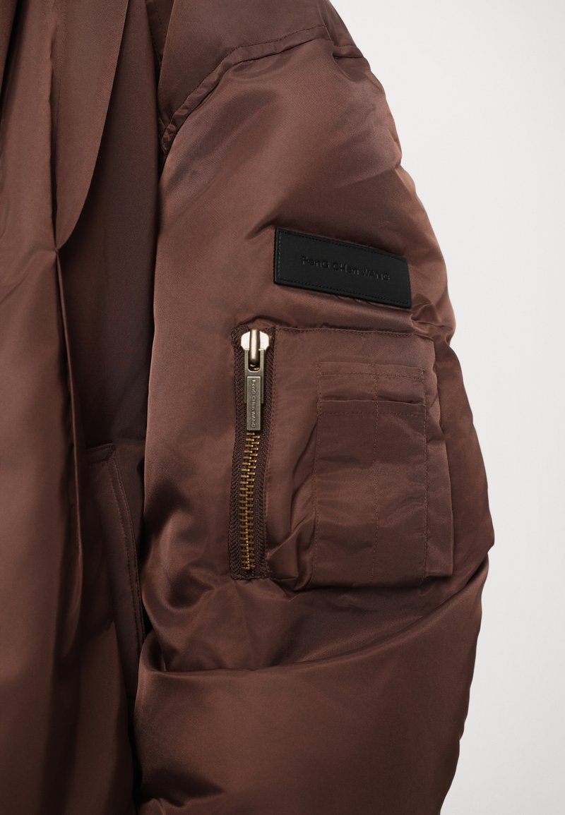 Braune Pufferjacke mit glattem, glänzendem Finish. Verfügt über eine aufgesetzte Tasche mit einem goldenen Reißverschluss und einem Etikett am Ärmel. Strukturierter Stoff.