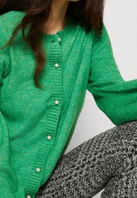 Zelený pletený cardigan s žebrovanou texturou a malými perlovými knoflíky, nošený přes vzorované černobílé legíny.