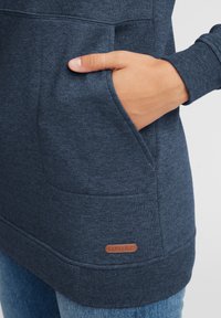 Mörkblå fleece-tröja med kenguruficka, ribbade muddar och en läderlogotyp på fållen. Slät textur och avslappnad passform.