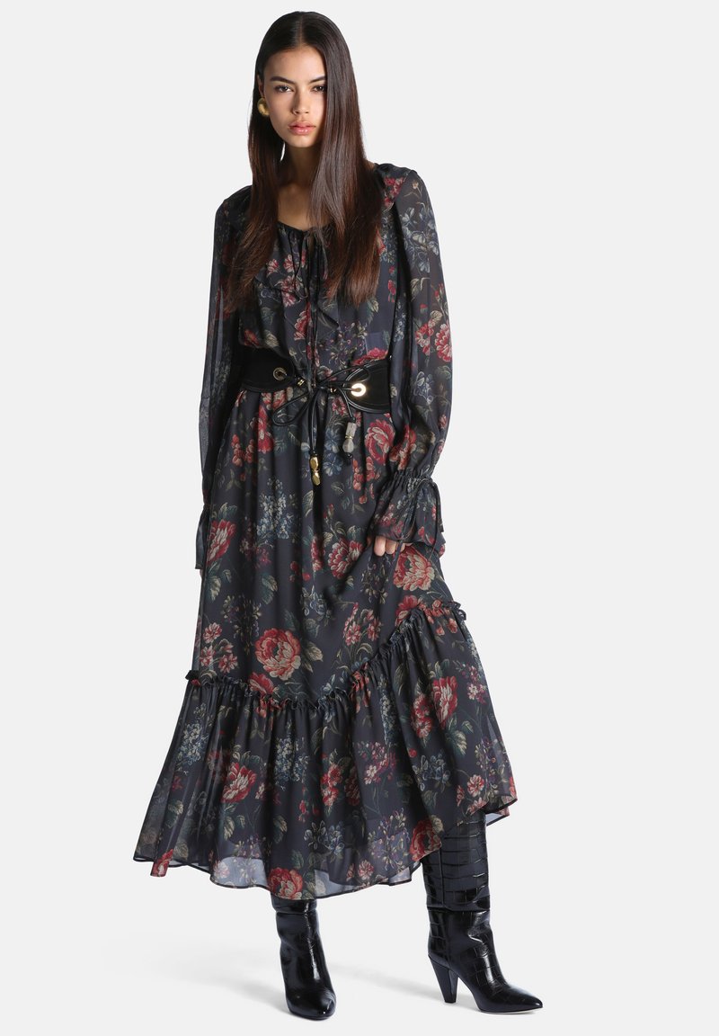 Femme portant une longue robe noire à fleurs avec des volants et une large ceinture noire, associée à des bottes à talons noires, debout face à l'avant.