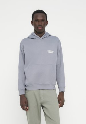 Homme portant un sweat à capuche violet clair avec le texte "Originals Studio" et un pantalon gris clair, debout devant un fond blanc uni.