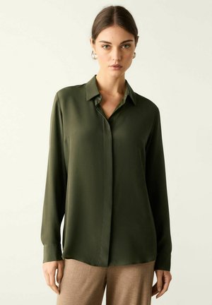 Frau, die eine dunkelgrüne langärmelige Bluse mit Knöpfen und beige Hosen trägt, steht vor einem einfarbigen hellen Hintergrund.