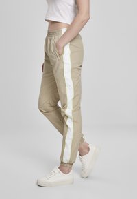 Beige treningsbukser med elastisk midje, sidelommer, hvite og neongrønne striper, avsmalnende mansjetter og myk, glatt tekstur.