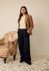 Bruine blazer met geborduurde accenten, crèmegebreide top en donkerblauwe wijde broekspijpen. Zwarte schoenen, bloemenbeklede stoel op de achtergrond.