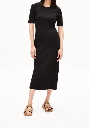 Kvinde iført en sort midi-kjole med korte ærmer og rund halsudskæring samt brune åbne sandaler, stående foran en ensartet hvid baggrund.