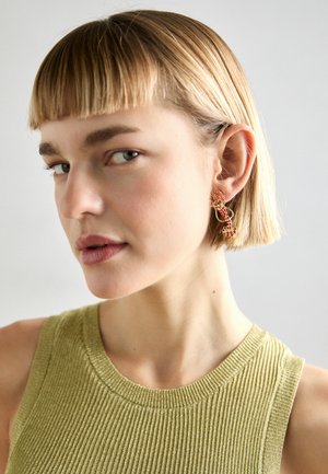 Goldfarbene Ohrringe mit roten dekorativen Akzenten und einem drehenden Design. Das Model trägt ein grünes, geripptes ärmelloses Oberteil und hat kurze Haare.