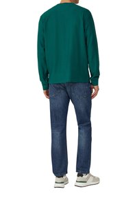 Zielona bluzka z długim rękawem o prążkowanej teksturze, w połączeniu z niebieskimi dżinsami i białymi trampkami. Bluzka ma okrągły dekolt i mankiety.