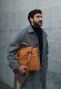 Man in gray herringbone coat holding a tan leather briefcase, standing against a gray concrete wall, looking to the right.