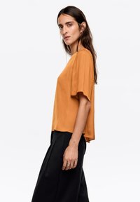 Krótki rękaw, top w kolorze rdzawego brązu z lekkiego materiału, z luźnym krojem i zaokrąglonym dołem, zestawiony z czarnymi spodniami o szerokich nogawkach.