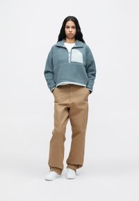 Γυναίκα με μακριά σκούρα μαλλιά, φορώντας γκρι φλις πουλόβερ, μπεζ παντελόνι σε φαρδιά γραμμή και λευκά αθλητικά παπούτσια, στέκεται με τα χέρια στην τσέπη.