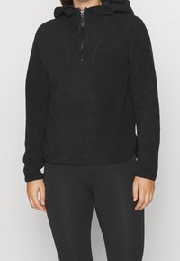 Czarna bluza polarowa z kapturem na zamek z przodu, luźnym krojem, sznurkiem przy kapturze i długimi rękawami, zestawiona z czarnymi legginsami. Miękka faktura.