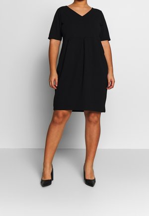 Kvinde iført en sort knælængde kjole med korte ærmer og V-udskæring, kombineret med sorte højhælede sko, stående på en ensartet baggrund.