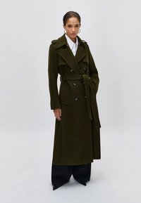 Olijfgroene dubbelgeknoopte trenchcoat met brede revers, een tailleband en grote knoppen, gecombineerd met wijde zwarte broek.