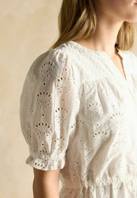 Weiße Baumwollbluse mit puffärmeln, die mit Lochstickerei und einem gerafften Bund versehen ist. Texturierter Stoff mit floralen Mustern und Rüschensäumen.