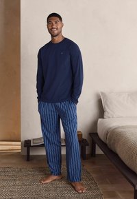 Tumši zilas krāsas krekls ar garām piedurknēm apvienots ar ziliem svītrainiem pidžamu biksēm. Audums izskatās mīksts, un bikses ir taisna griezuma ar elastīgu jostu.