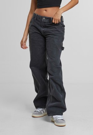 Person iført løse sorte denim bukser med værktøjslomme og grå sneakers, som holder bukselinningen med den ene hånd.