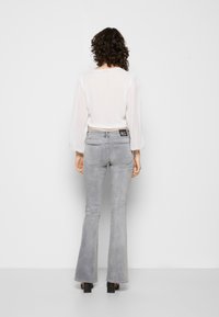 Jeans cinzentos com forma flare e um ligeiro elastano, apresentando uma cintura de altura média e dois bolsos traseiros. Combinados com uma blusa branca, transparente, de mangas compridas.