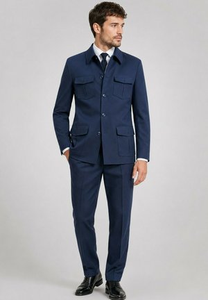 Homme portant un costume bleu marine avec une veste boutonnée à quatre poches, une chemise blanche, une cravate noire et des chaussures habillées noires, debout les mains dans les poches.