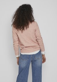 Ljusrosa tröja med ribbade ärmslut och nederkant, lager-på-lager över en vit skjorta, tillsammans med vida blå jeans. Mjuk textur, avslappnad passform.