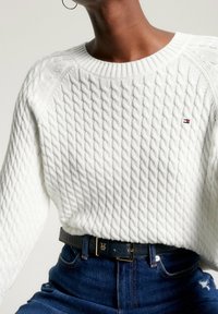 Witte kabelgebreide trui met een geribde halslijn en een gestructureerd patroon, gecombineerd met blauwe denim jeans en een zwarte riem met een gouden gesp.